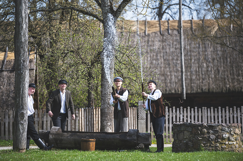 Lany Poniedziałek. Scena plenerowa z muzeum na wolnym powietrzu. Czterech mężczyzn w tradycyjnych strojach (czapki maciejówki, kamizelki) wylewa dynamiczne strumienie wody z długich drewnianych rur (dyngusówki). Akcja toczy się przy starej studni i chatach krytych strzechą.