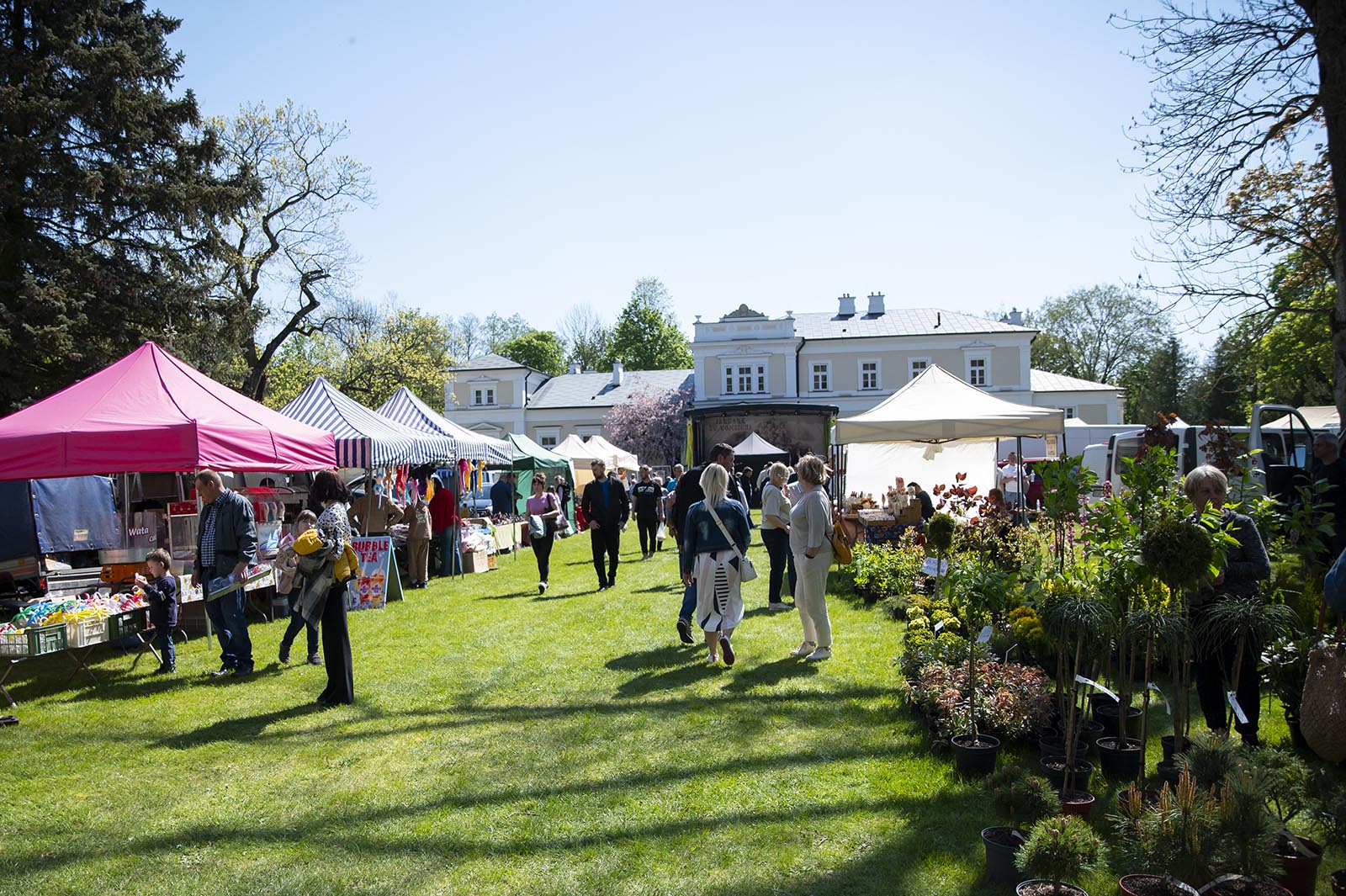 Tłum ludzi spaceruje pomiędzy kolorowymi namiotami handlowymi podczas plenerowego jarmarku. Po lewej stronie znajdują się stoiska z różnymi towarami, w tym różowy i pasiaste namioty, a po prawej duże stoisko oferujące rośliny i krzewy w doniczkach. W słonecznym tle, za zielonym trawnikiem, widoczny jest jasny budynek o architekturze dworkowej otoczony wysokimi drzewami.