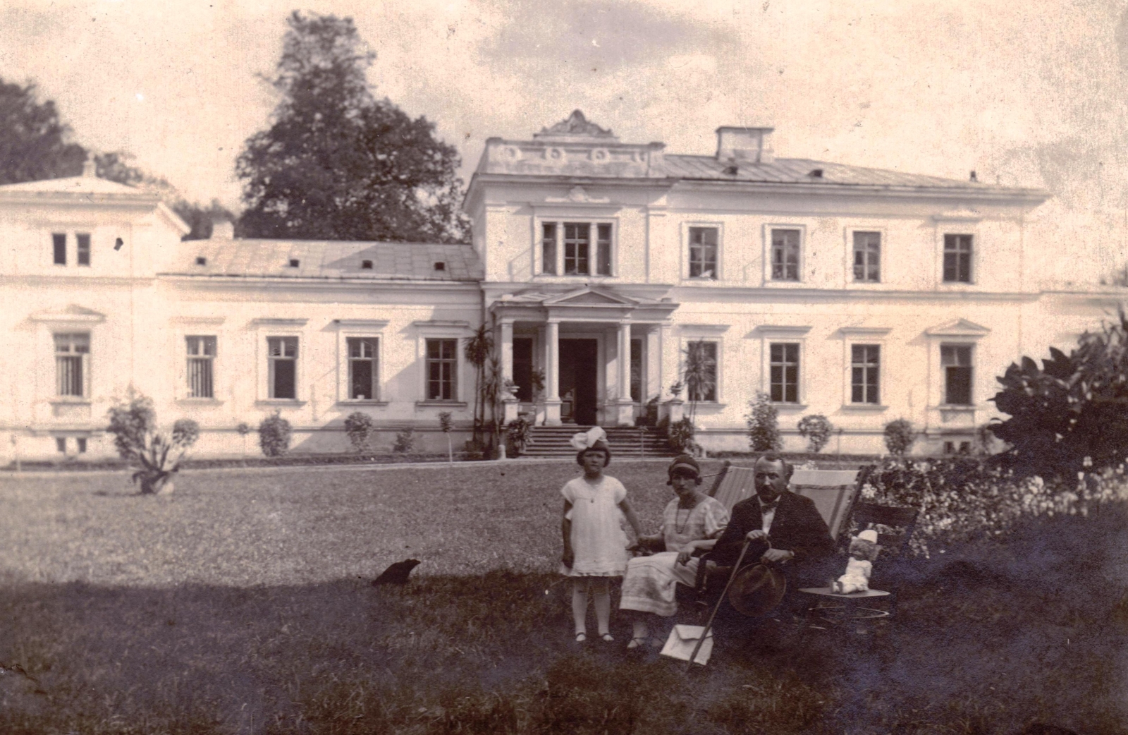 Czarno-biała, archiwalna fotografia przedstawiająca okazały dwór z kolumnowym wejściem. Na trawniku przed budynkiem siedzi mężczyzna z laską, obok niego kobieta i mała dziewczynka w jasnej sukience.