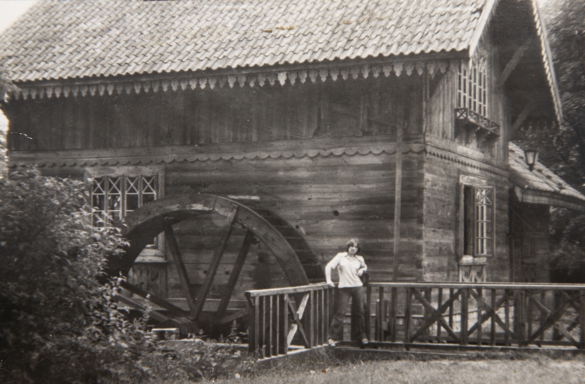 Archiwalne czarno-białe zdjęcie drewnianego młyna z dużym kołem wodnym. Na drewnianym pomoście przed budynkiem stoi młoda osoba oparta o balustradę.