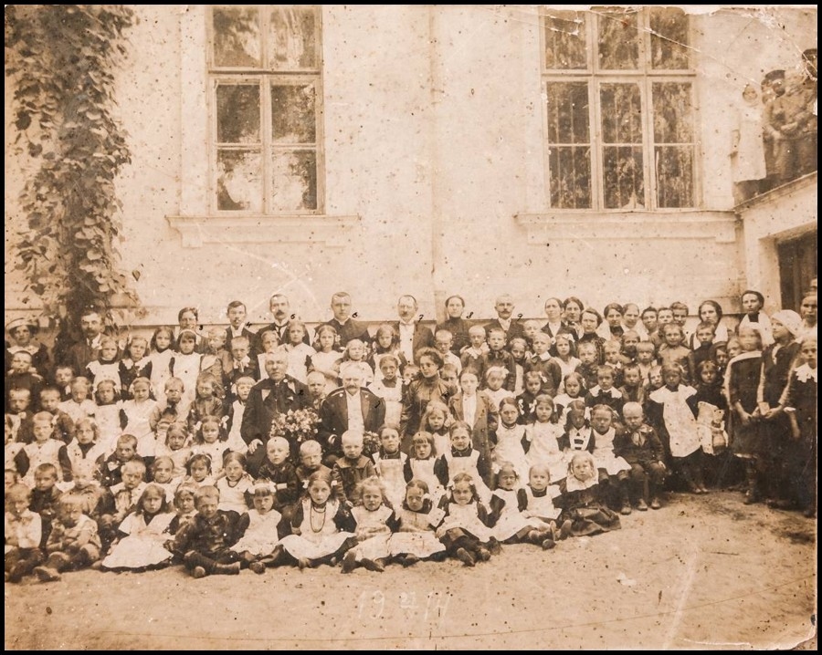 Stara, zbiorowa fotografia grupy dzieci i dorosłych ustawionych przed budynkiem. W pierwszym rzędzie siedzą dzieci, za nimi stoją opiekunowie; zdjęcie datowane na 1914 rok.