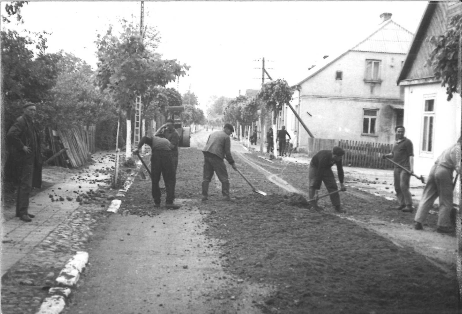 Czarno-białe zdjęcie przedstawiające mężczyzn pracujących przy naprawie lub budowie drogi w małej miejscowości. W tle widoczne domy, ogrodzenia oraz przechodnie.