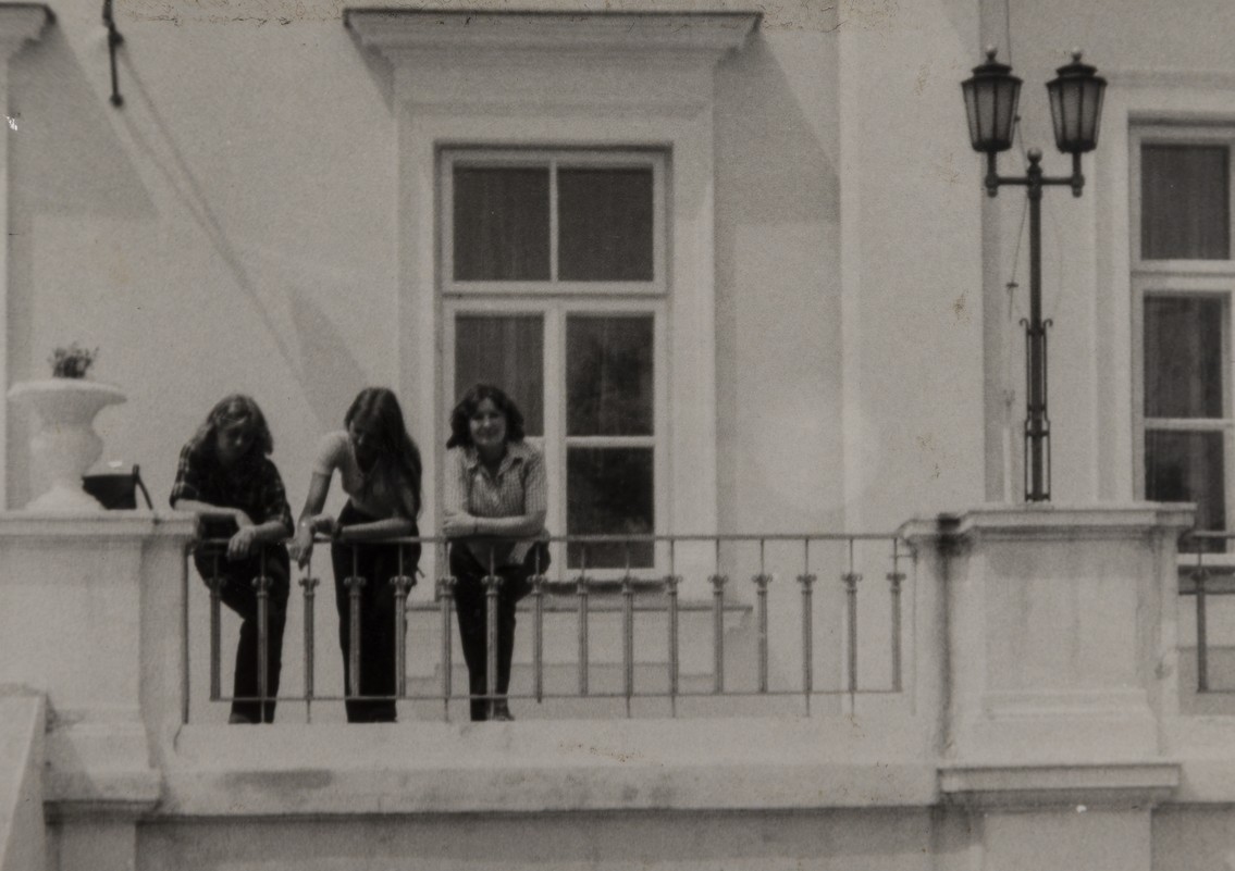 Czarno-białe zdjęcie trzech młodych kobiet stojących na balkonie jasnego budynku. Kobiety opierają się o metalową balustradę i patrzą w dół.