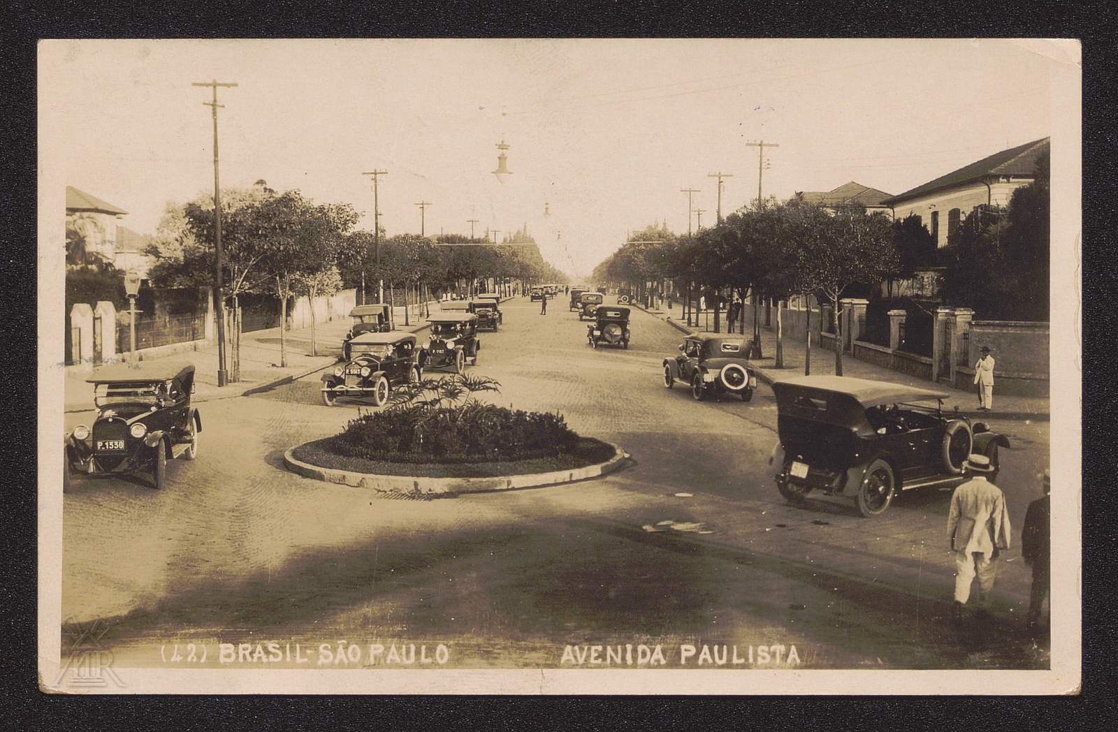 Archiwalna fotografia przedstawiająca Avenida Paulista w São Paulo w Brazylii. Szeroka, brukowana aleja z zabytkowymi samochodami, szpalerami drzew i zabudową miejską.