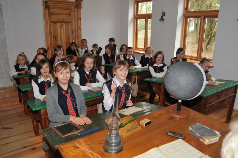 Klasa z uczniami w mundurkach siedzącymi przy drewnianych ławkach z zielonymi blatami. Na stole stoi globus. W pomieszczeniu jest jasno, dzięki naturalnemu oświetleniu.