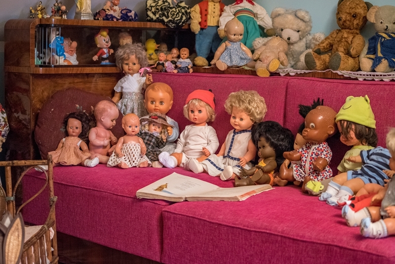 Kolekcja zabytkowych lalek ustawiona jest na różowej sofie, otoczona pluszakami. Przed nią leży otwarta książka, tworząc nostalgiczną atmosferę.