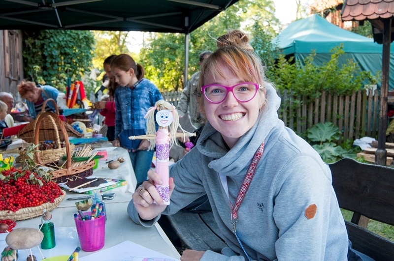 Uśmiechnięta kobieta w różowych okularach trzyma ręcznie wykonaną lalkę przy stoliku z artykułami rękodzielniczymi na świeżym powietrzu, otoczona materiałami artystycznymi i kolorowymi koszami.