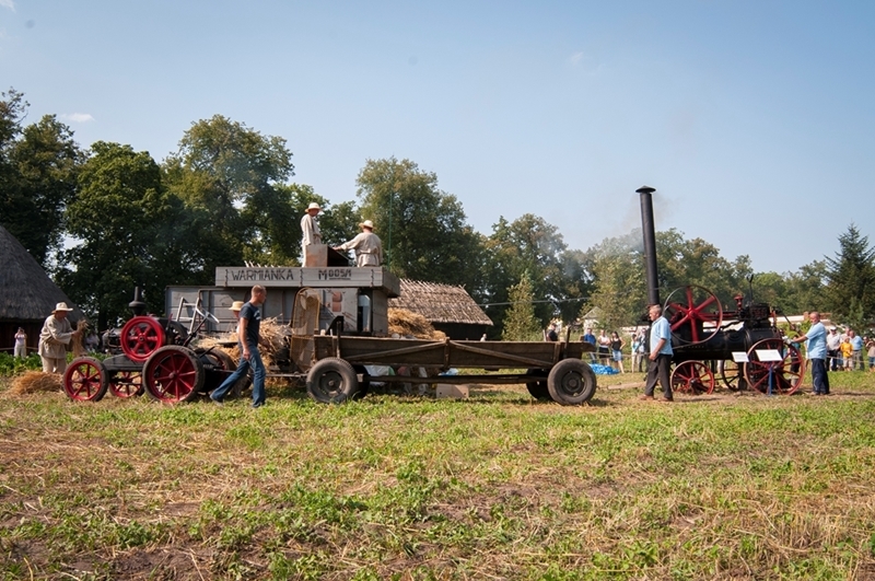 Scena z farmy z ludźmi obsługującymi maszynę parową i młocarnię na trawiastym polu. Atmosfera jest nostalgiczna i żywa.