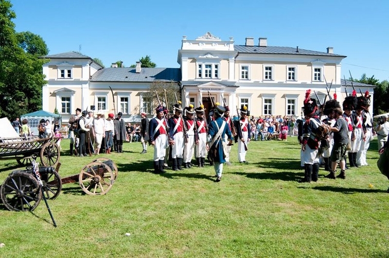 Rekonstrukcja historyczna na trawiastym terenie, z ludźmi w historycznych mundurach wojskowych ustawionymi w dwóch rzędach. Widoczny jest zabytkowy powóz i duży tłum, a w tle – majestatyczny budynek pod czystym, błękitnym niebem – tworzy to atmosferę nostalgii i historycznej refleksji.