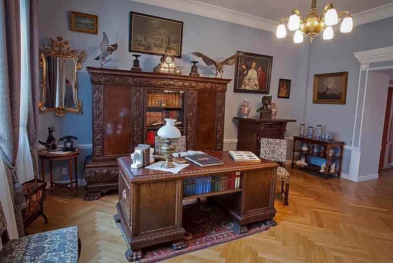 Elegancki gabinet z ozdobnym drewnianym biurkiem i regałami na książki, ozdobiony antycznymi dekoracjami, obrazami i żyrandolem. Atmosfera jest klasyczna i wyrafinowana.