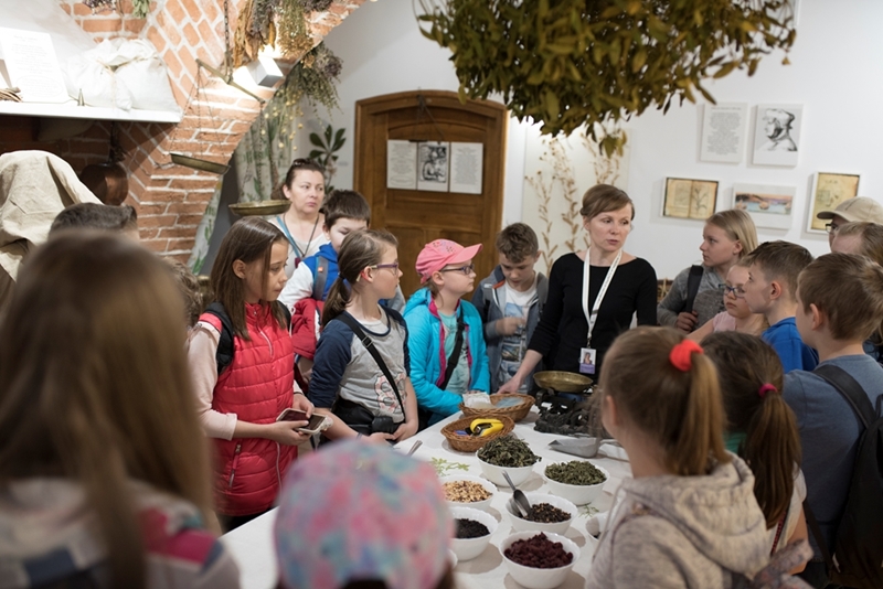 Grupa dzieci gromadzi się wokół stołu z miskami ziół, podczas gdy kobieta coś wyjaśnia. Przytulne otoczenie, z ceglanym łukiem i roślinami wiszącymi nad nim.
