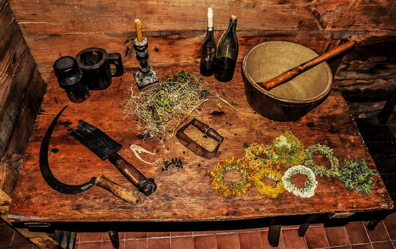 Rustykalny, drewniany stół z suszonymi ziołami, dwiema butelkami, zapaloną świecą i zabytkowymi narzędziami kuchennymi. Ciepłe oświetlenie nadaje wnętrzu przytulny, historyczny charakter.