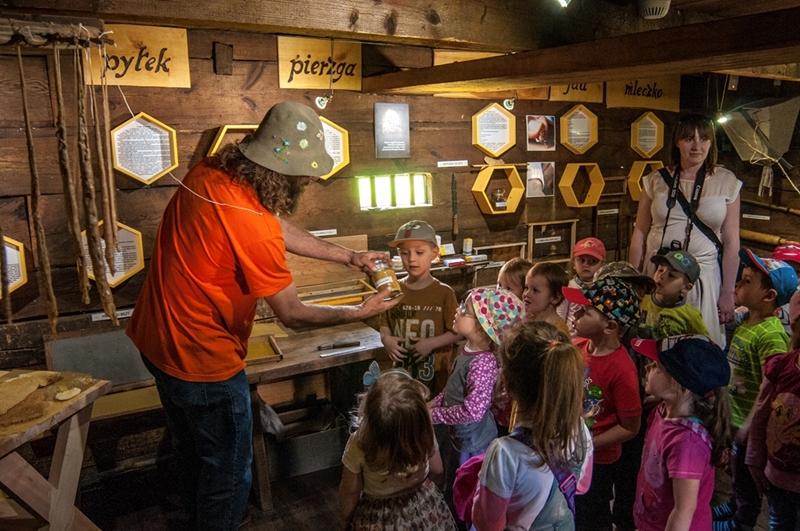 Mężczyzna w pomarańczowej koszuli pokazuje słoik grupie ciekawskich dzieci w przytulnym, drewnianym muzeum. Atmosfera jest edukacyjna i angażująca.