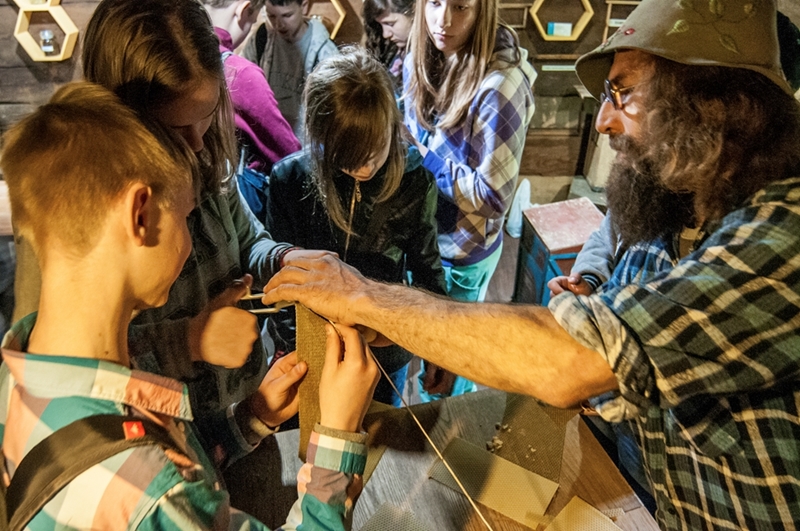 Brodaty mężczyzna w kapeluszu uczy dzieci, jak zrobić świece z wosku pszczelego. Dzieci są skupione i zainteresowane, co tworzy ciepłą, edukacyjną atmosferę.