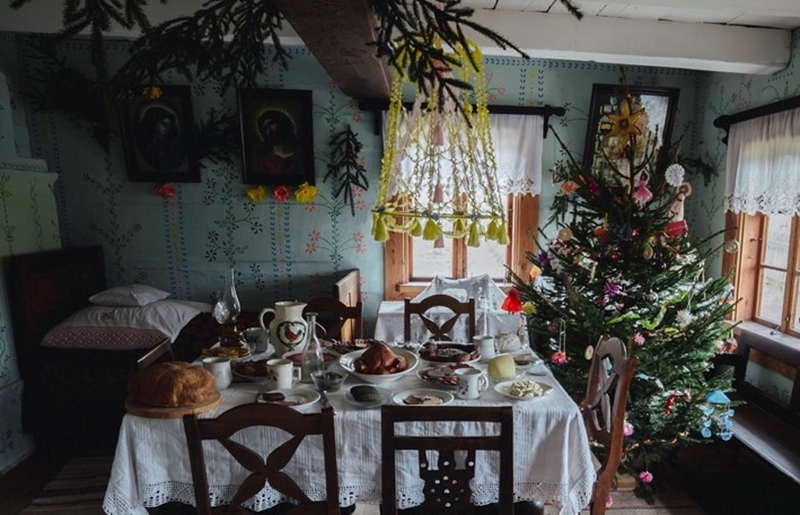 Przytulna jadalnia ze świątecznym stołem nakrytym białym obrusem, tradycyjnymi potrawami i udekorowaną choinką w pobliżu.