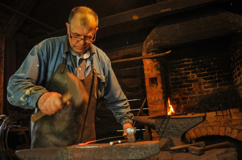 Kowal ubrany w niebieską koszulę i fartuch wykuwa metal na kowadle w słabo oświetlonym warsztacie, w pobliżu tli się ogień w ceglanym piecu.