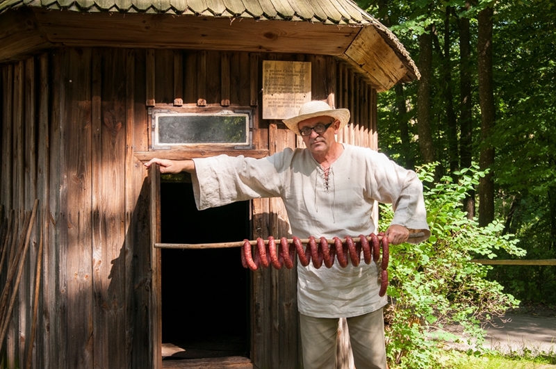 Osoba w tradycyjnym stroju i słomkowym kapeluszu stoi przed drewnianą chatą, trzymając długi kij z wieloma kiełbaskami. Scena jest rustykalna i spokojna.