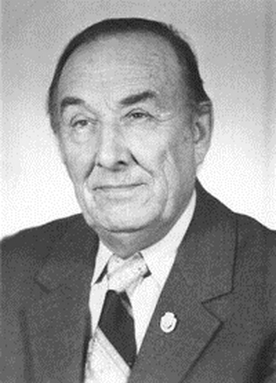 Starszy mężczyzna w garniturze i krawacie, o spokojnym wyrazie twarzy. W klapie jego marynarki znajduje się mała szpilka. Czarno-białe zdjęcie oddaje poczucie godności i historii.