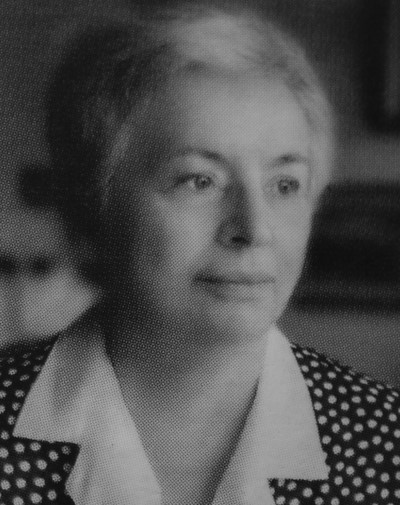 Czarno-białe zdjęcie starszej kobiety z krótkimi włosami, ubranej w sukienkę w groszki. Spogląda zamyślona w bok, co emanuje spokojnym, refleksyjnym nastrojem.