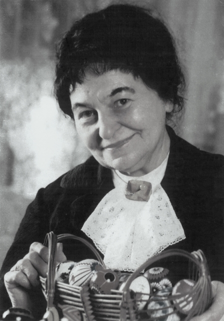 Starsza kobieta o ciepłym uśmiechu trzyma pleciony kosz pełen kolorowych guzików. Ma na sobie bluzkę z koronkowym kołnierzykiem i ciemną marynarkę.