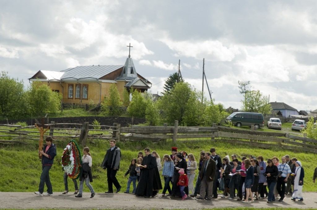 Procesja ludzi idzie wiejską drogą, prowadzona przez osobę trzymającą krzyż i wieniec. W tle, na tle częściowo zachmurzonego nieba, widać mały kościółek z krzyżem na dachu. Scena ta emanuje uroczystą, a zarazem spokojną atmosferą.