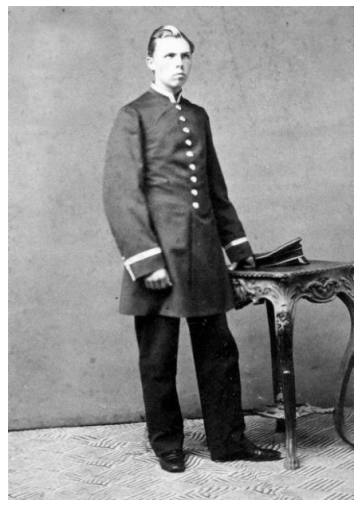 Czarno-białe zdjęcie mężczyzny w oficjalnym mundurze wojskowym z mosiężnymi guzikami, stojącego przy ozdobnym stole. Wygląda na poważnego i opanowanego.