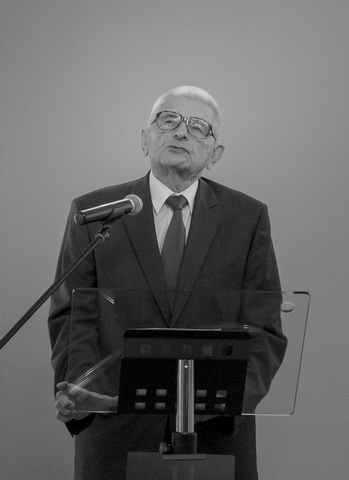 Starszy mężczyzna w garniturze i krawacie przemawia z zamyśloną miną na podium. Obok niego stoi mikrofon. 