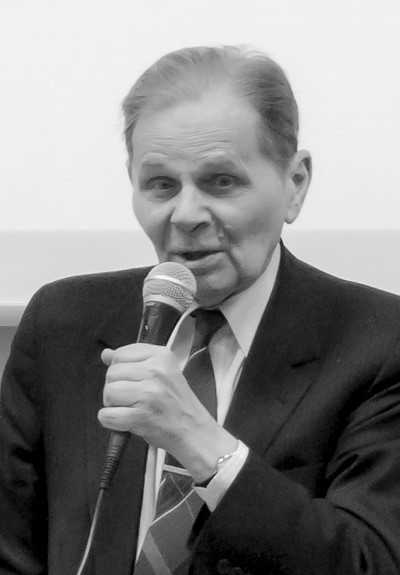 Starszy mężczyzna w garniturze mówi do mikrofonu. Czarno-biały obraz o formalnym tonie, prawdopodobnie podczas wykładu lub prezentacji.