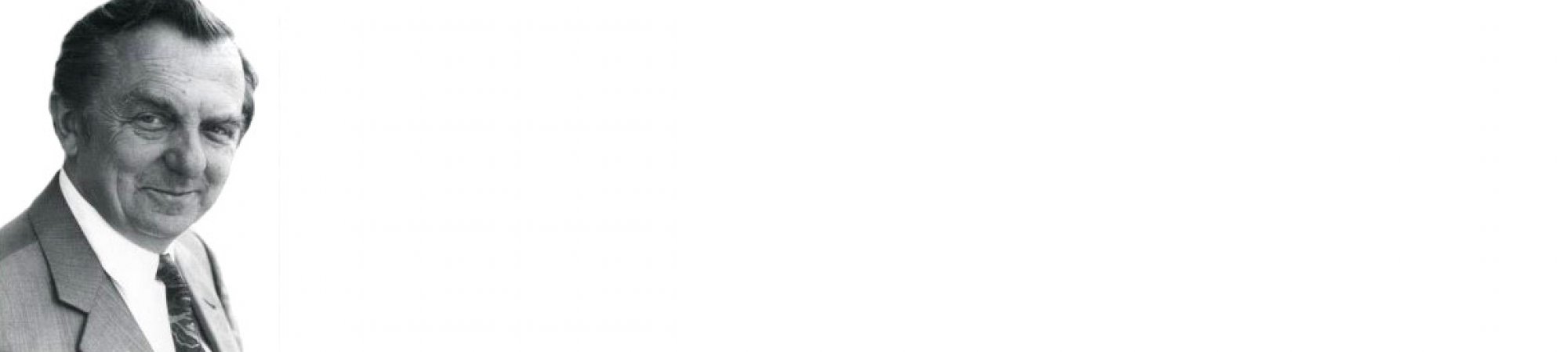 Starszy mężczyzna w garniturze i krawacie, uśmiechając się subtelnie, stoi na białym tle.