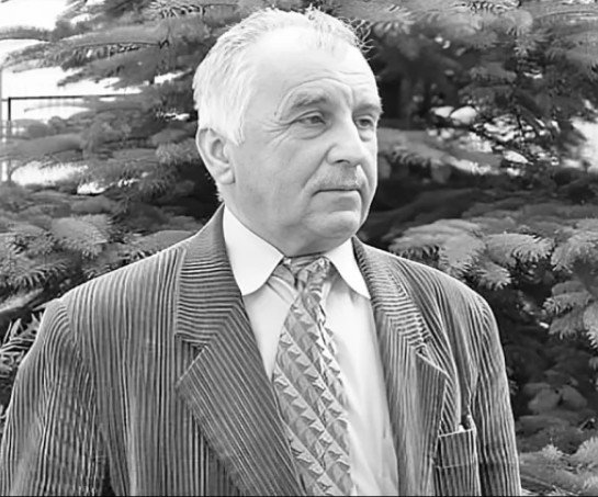 Starszy mężczyzna w garniturze i wzorzystym krawacie stoi zamyślony na zewnątrz, na tle sosny. Czarno-białe zdjęcie emanuje nostalgicznym tonem.