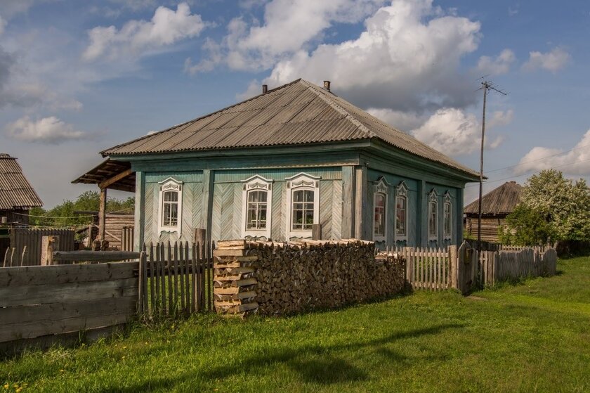 Urokliwy drewniany dom z turkusowymi ścianami i szarym, spadzistym dachem otoczony jest drewnianym płotem. Na zewnątrz starannie ułożone są kłody drewna. Błękitne niebo z puszystymi chmurami tworzy spokojną, wiejską atmosferę.