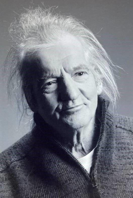 Starsza osoba z długimi, nieuczesanymi, białymi włosami i łagodnym wyrazem twarzy, ubrana w rozpinany sweter na spokojnym portrecie w odcieniach szarości.