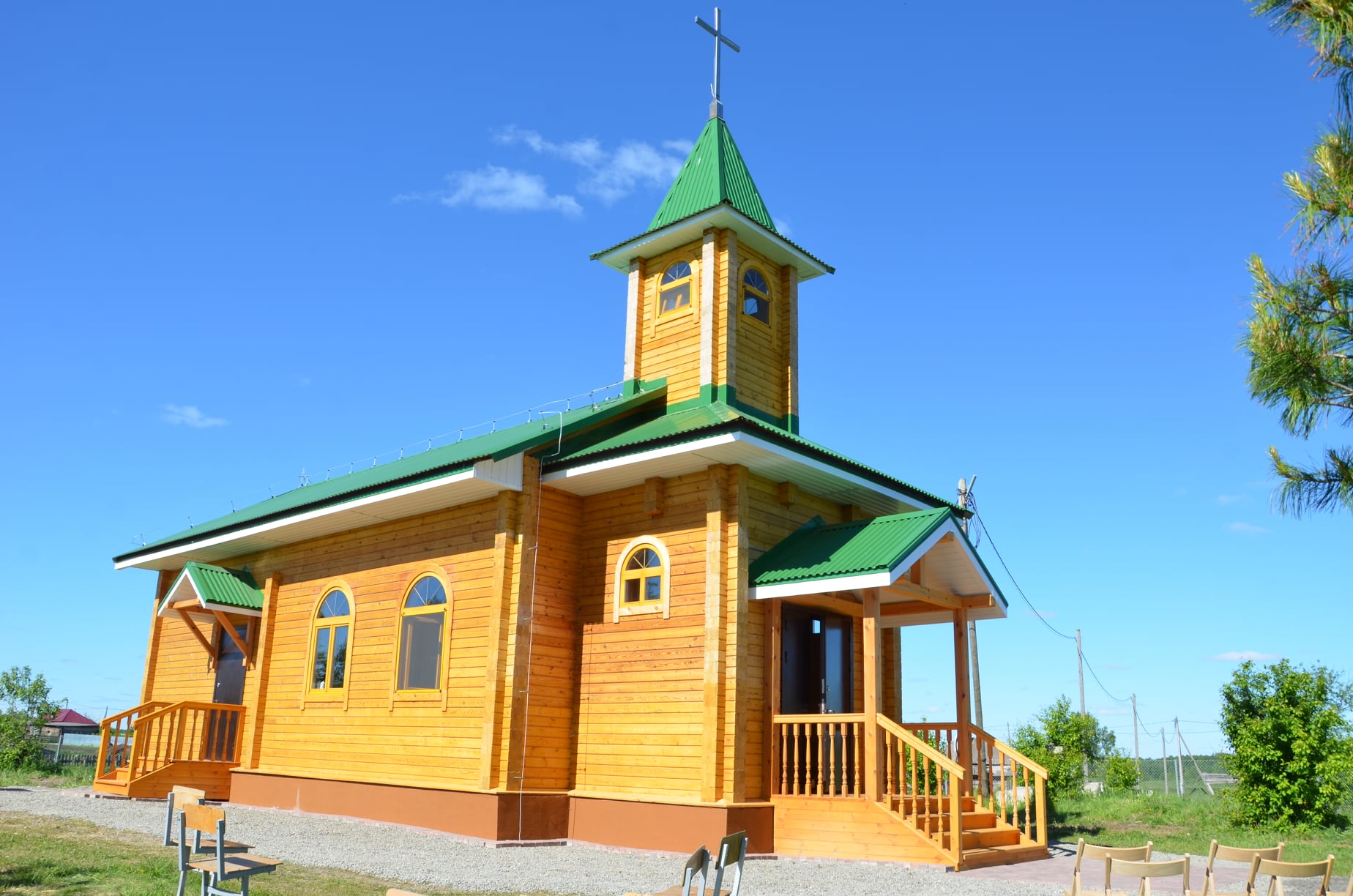 Mały drewniany kościółek z zielonymi dachami stoi pod czystym, błękitnym niebem. Jego łukowate okna i krzyż na szczycie wieży tworzą atmosferę spokoju.